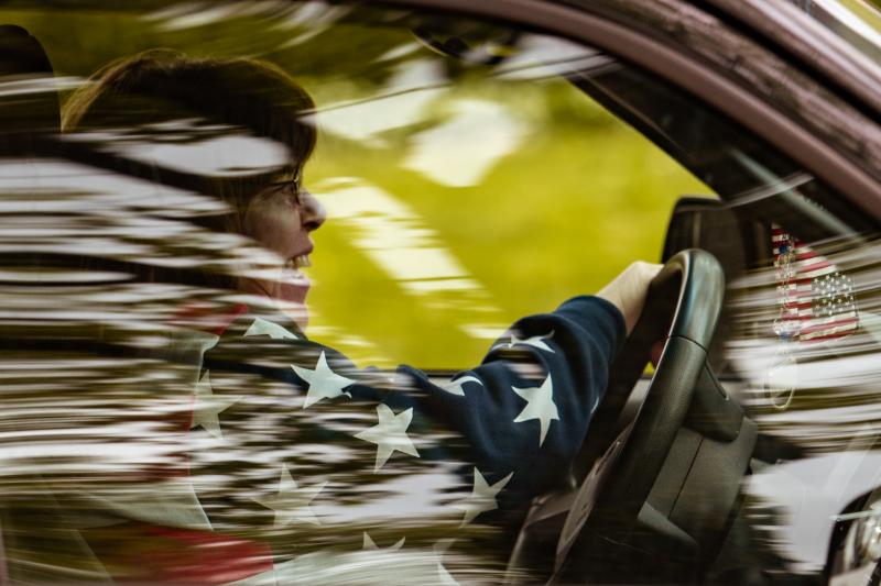 Person sits at steering wheel of car in an American flag jacket, with motion blur