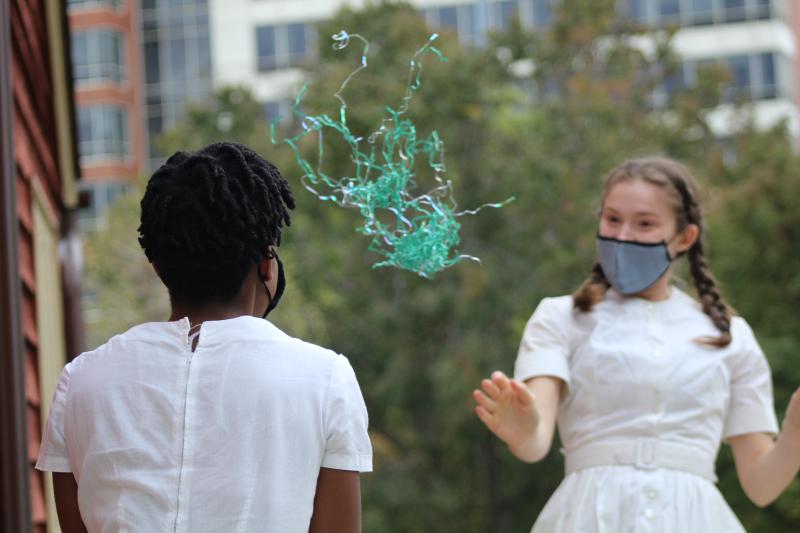 Masked woman in white dress tosses green blobby thing to other masked woman in white dress