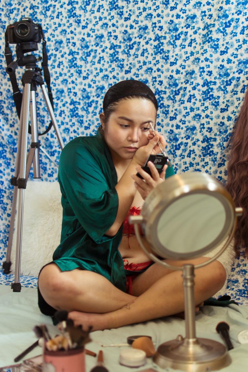 Sex worker applies make-up in front of hand-held mirror with tripod in background