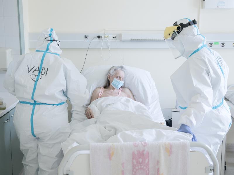 Two doctors in white full-body hazmat suits stand on either side of a patient's hospital bed