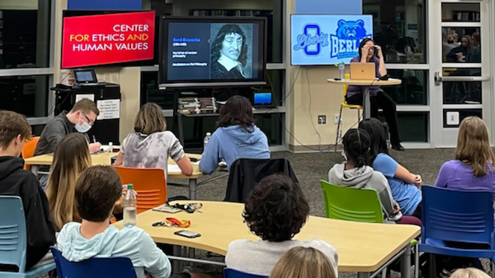Backs of students seated at desks and listening to Lisa Shabel speak on Descartes, whose image appears on a projector screen, as does a CEHV logo. 