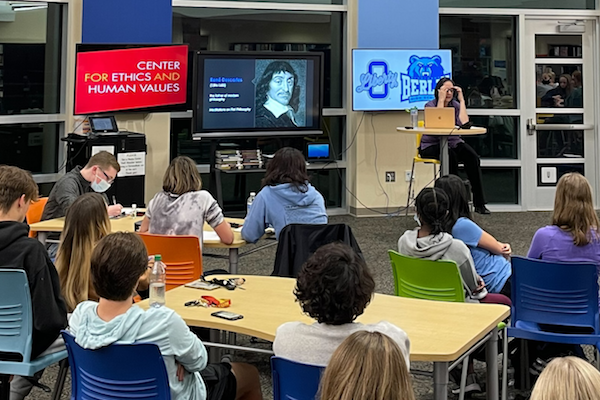 Backs of students seated at desks and listening to Lisa Shabel speak on Descartes, whose image appears on a projector screen, as does a CEHV logo. 
