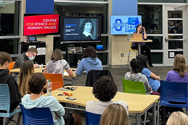 Backs of students seated at desks and listening to Lisa Shabel speak on Descartes, whose image appears on a projector screen, as does a CEHV logo.