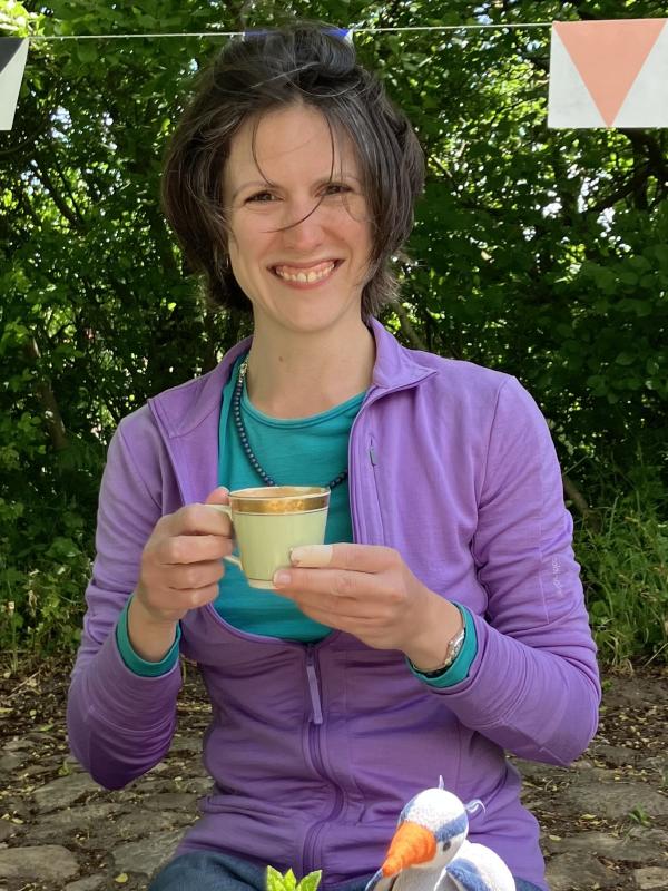 Kate McFarland sitting outside in front of some trees smiling and holding a coffee cup.